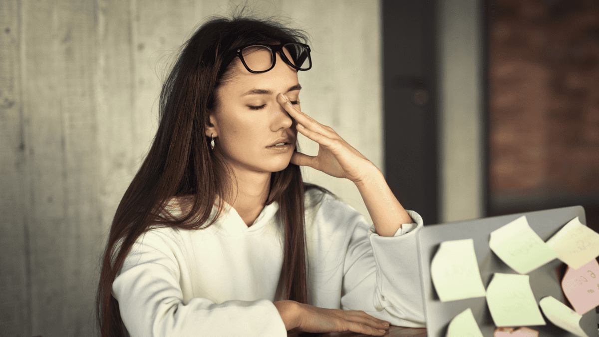 A woman sitting at a desk with a laptop, holding her forehead with one hand while looking stressed, with glasses resting on her head and sticky notes scattered on the laptop.
