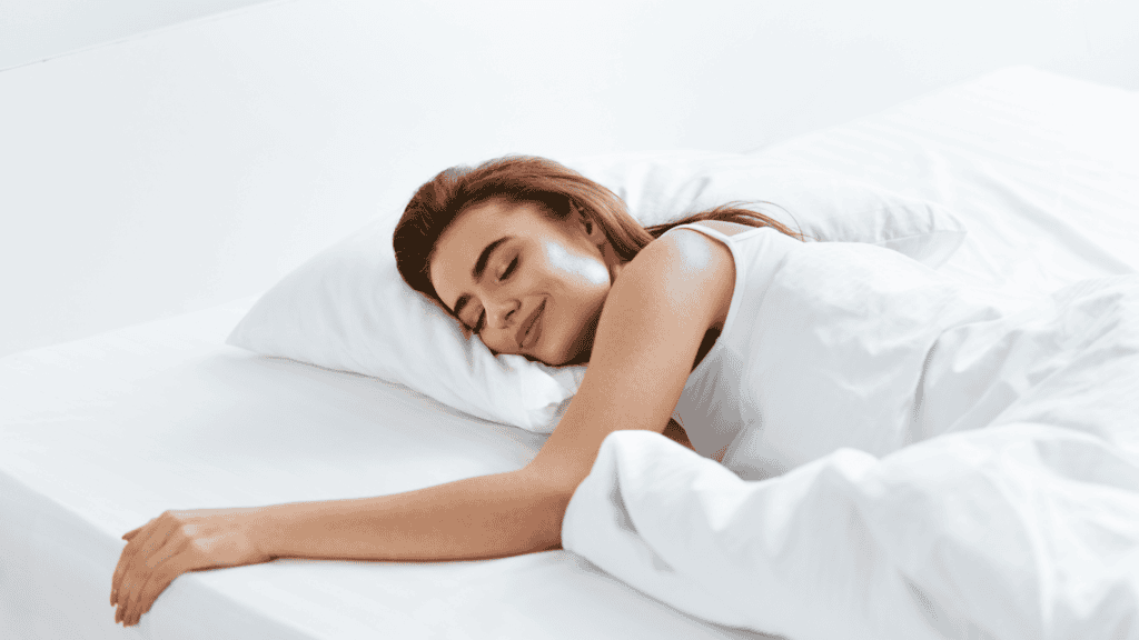 A woman peacefully sleeping in a bed, smiling while lying on her side with her arm resting on the pillow and covered with a white blanket.