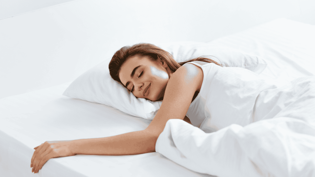 A woman peacefully sleeping in a bed, smiling while lying on her side with her arm resting on the pillow and covered with a white blanket.