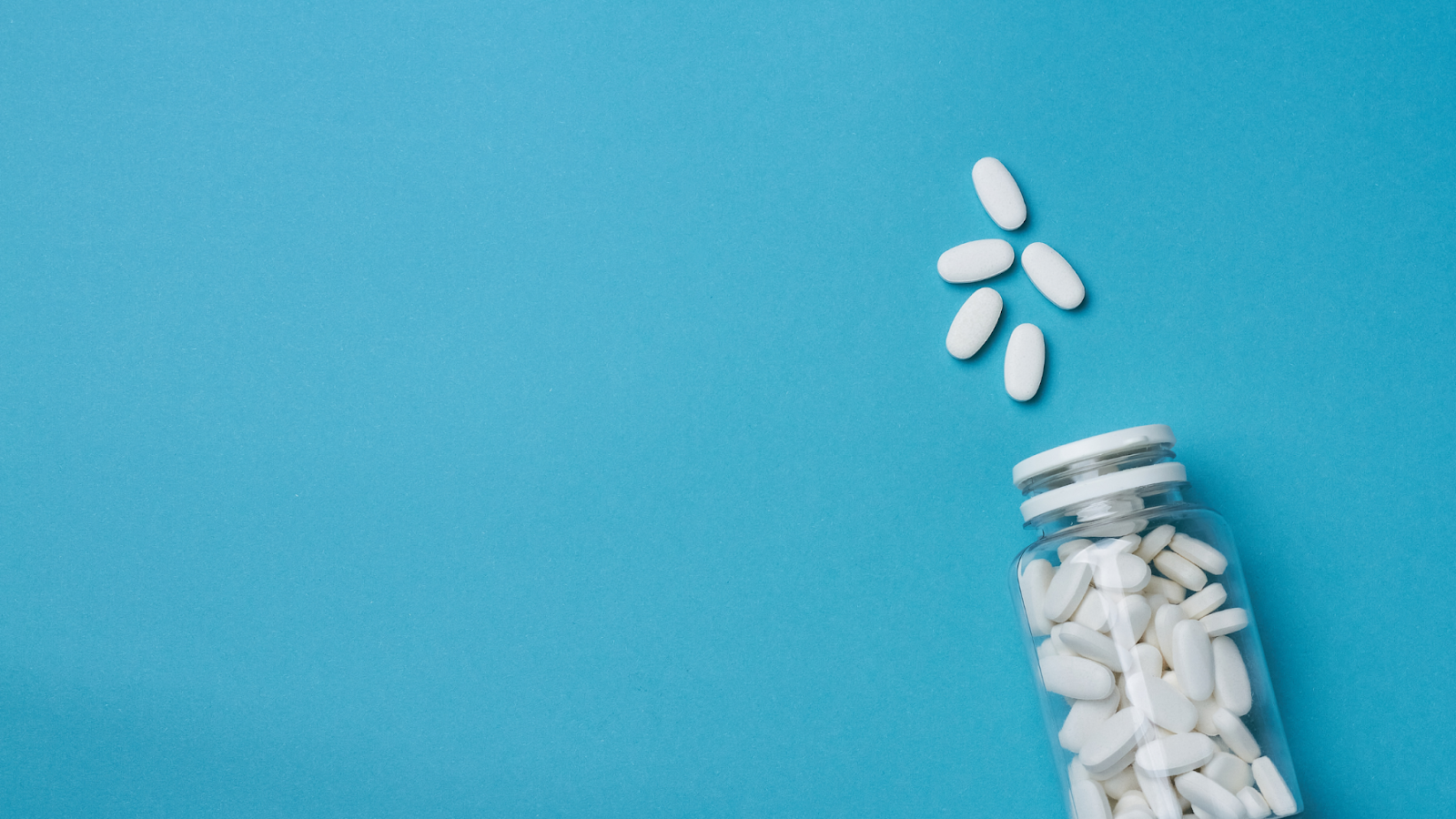 White oval pills spilling from a clear bottle on blue background