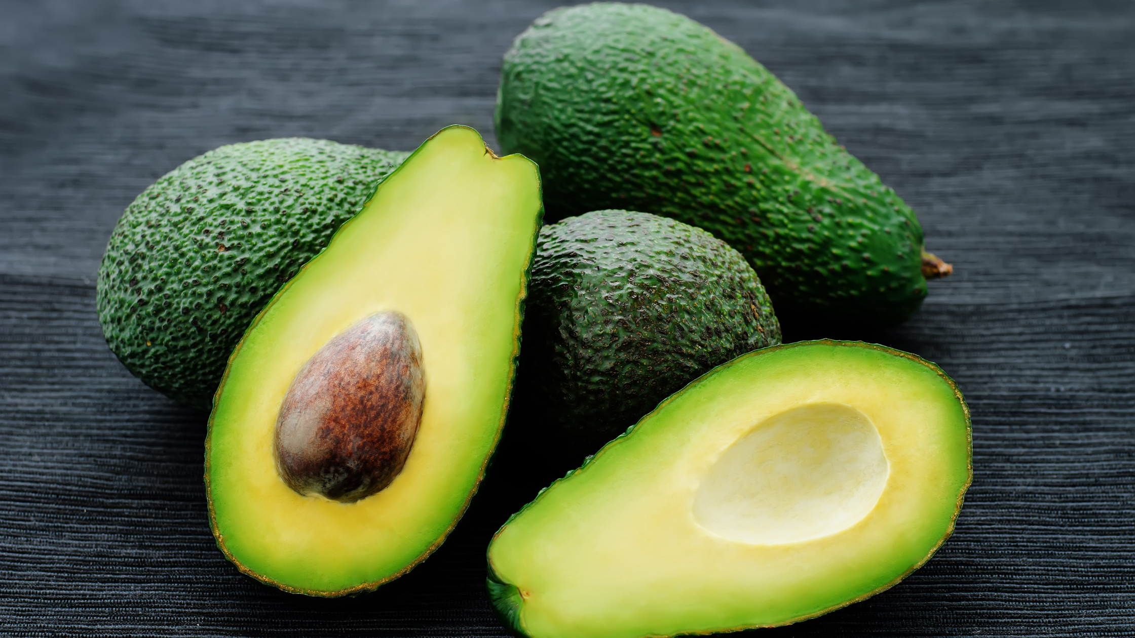 Whole and halved ripe avocados on dark surface