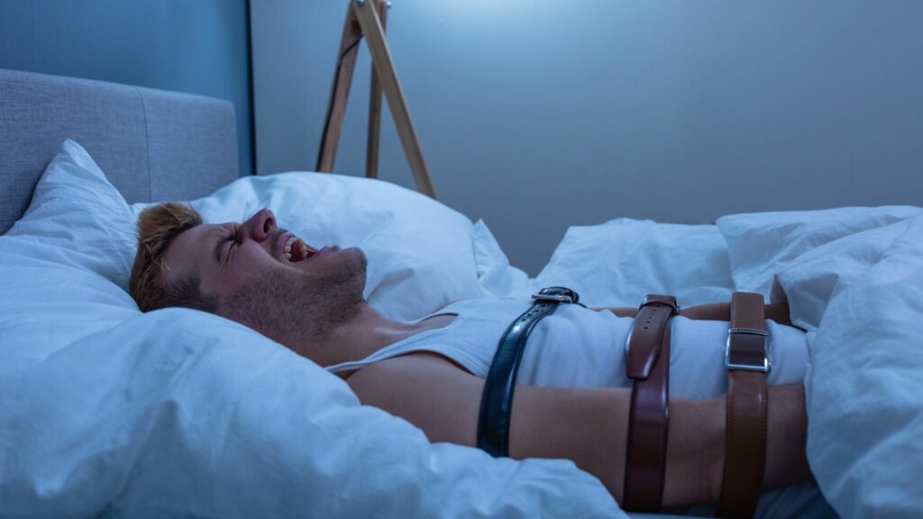 Illustration showing a person lying awake in bed, unable to move, with a shadowy figure nearby, depicting common sleep paralysis symptoms.