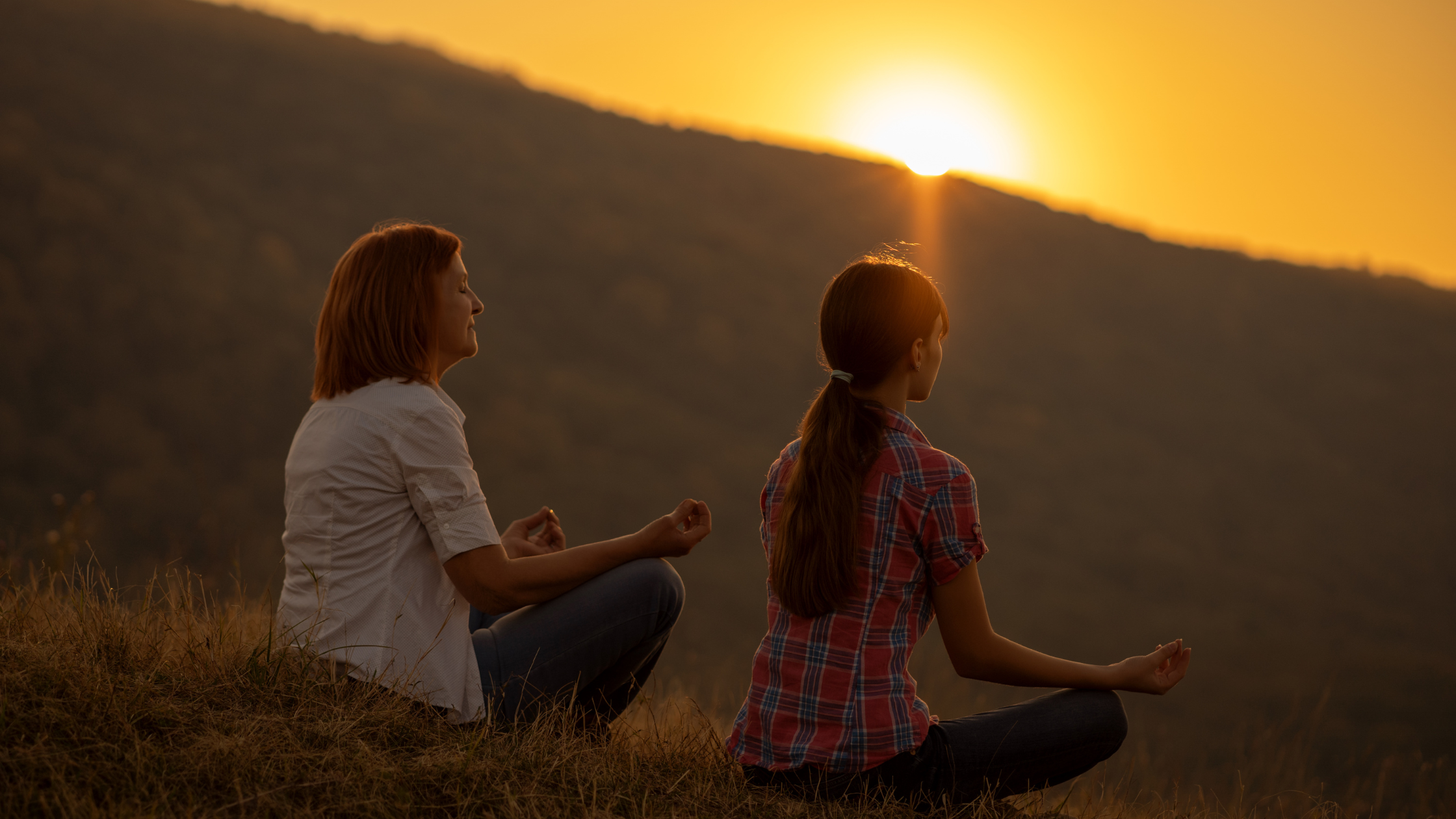Two women meditating outdoors at sunset on a grassy hill