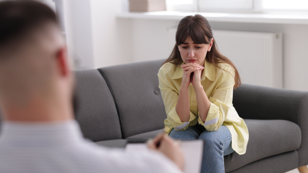 A person sits on a couch holding a tissue, looking thoughtful and sad, while a therapist offers supportive guidance during a breakup therapy session.