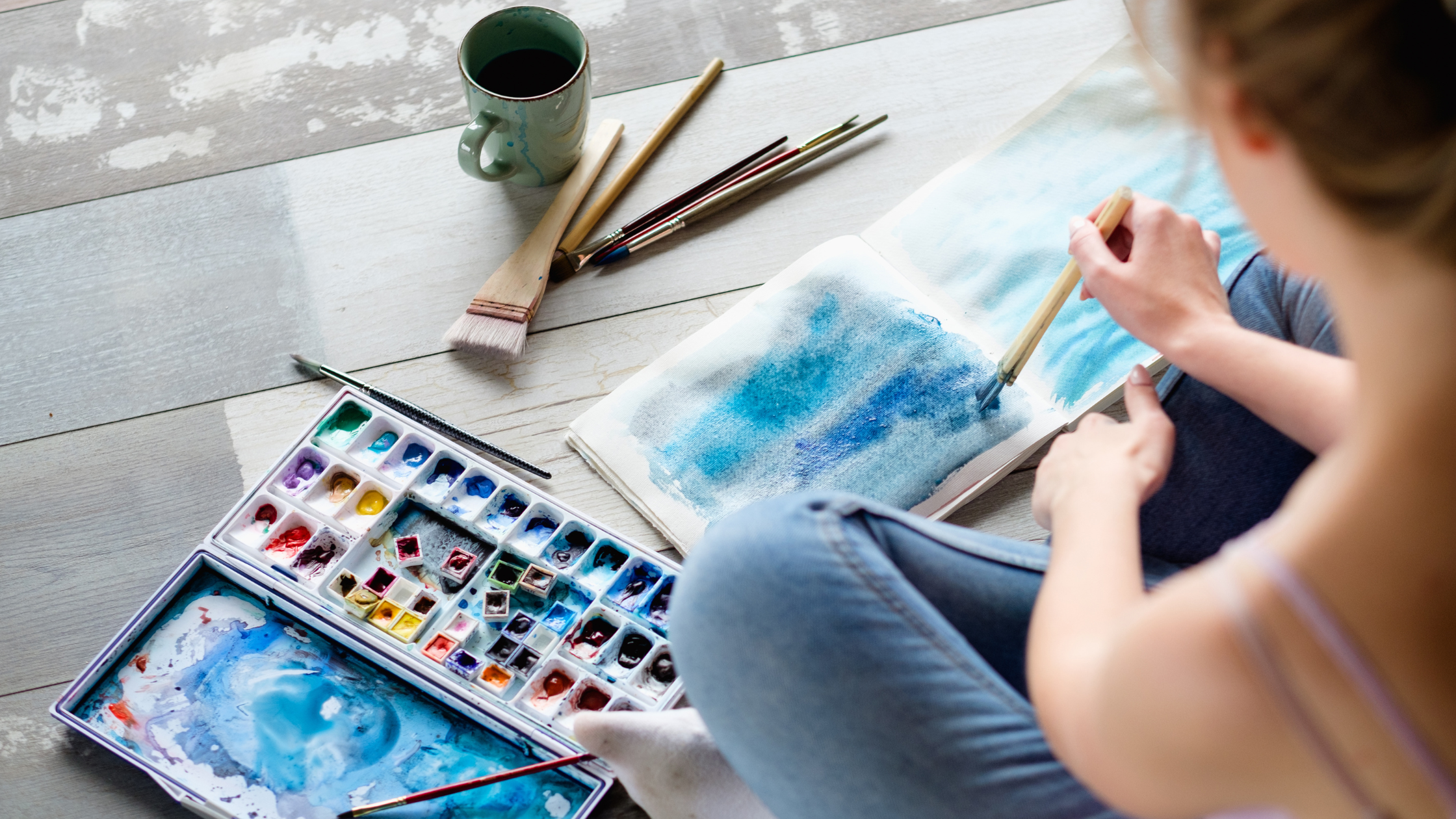 Person painting blue watercolor in sketchbook on floor