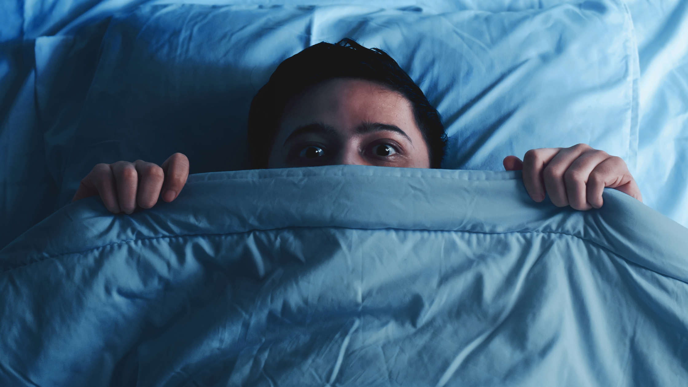 Person hiding under blue blanket with wide scared eyes