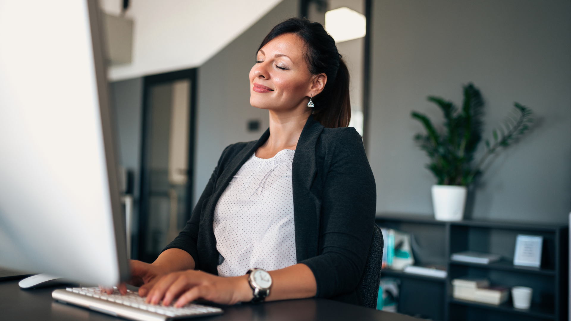 Smiling employee sitting at a desk with a laptop, surrounded by plants and personal items, showing how to be happy and comfortable at work.