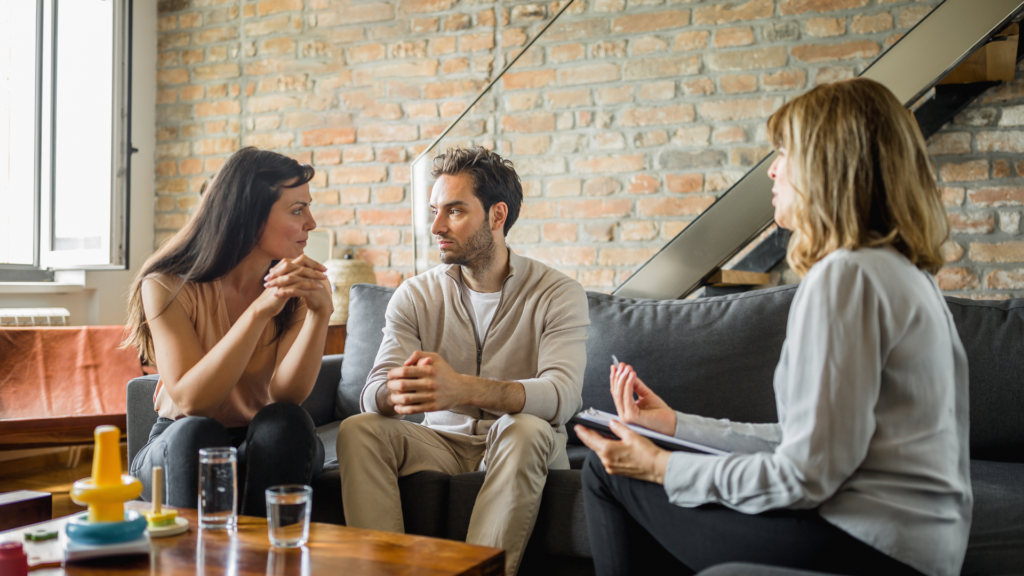 A couple sits on a couch holding hands, talking with a therapist about managing anxiety together during a couples therapy session.