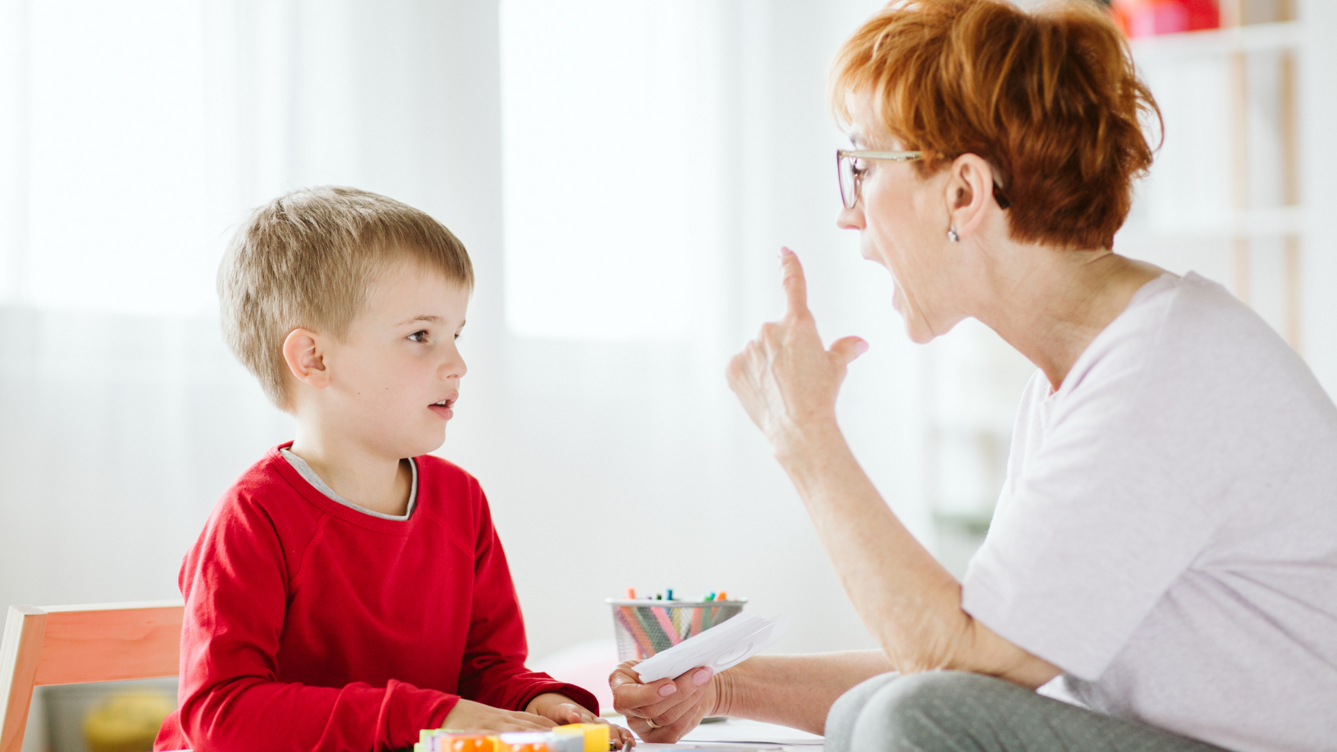 A young child sits quietly with a thoughtful expression, while a caregiver gently encourages speech, illustrating signs of speech delay in early childhood.