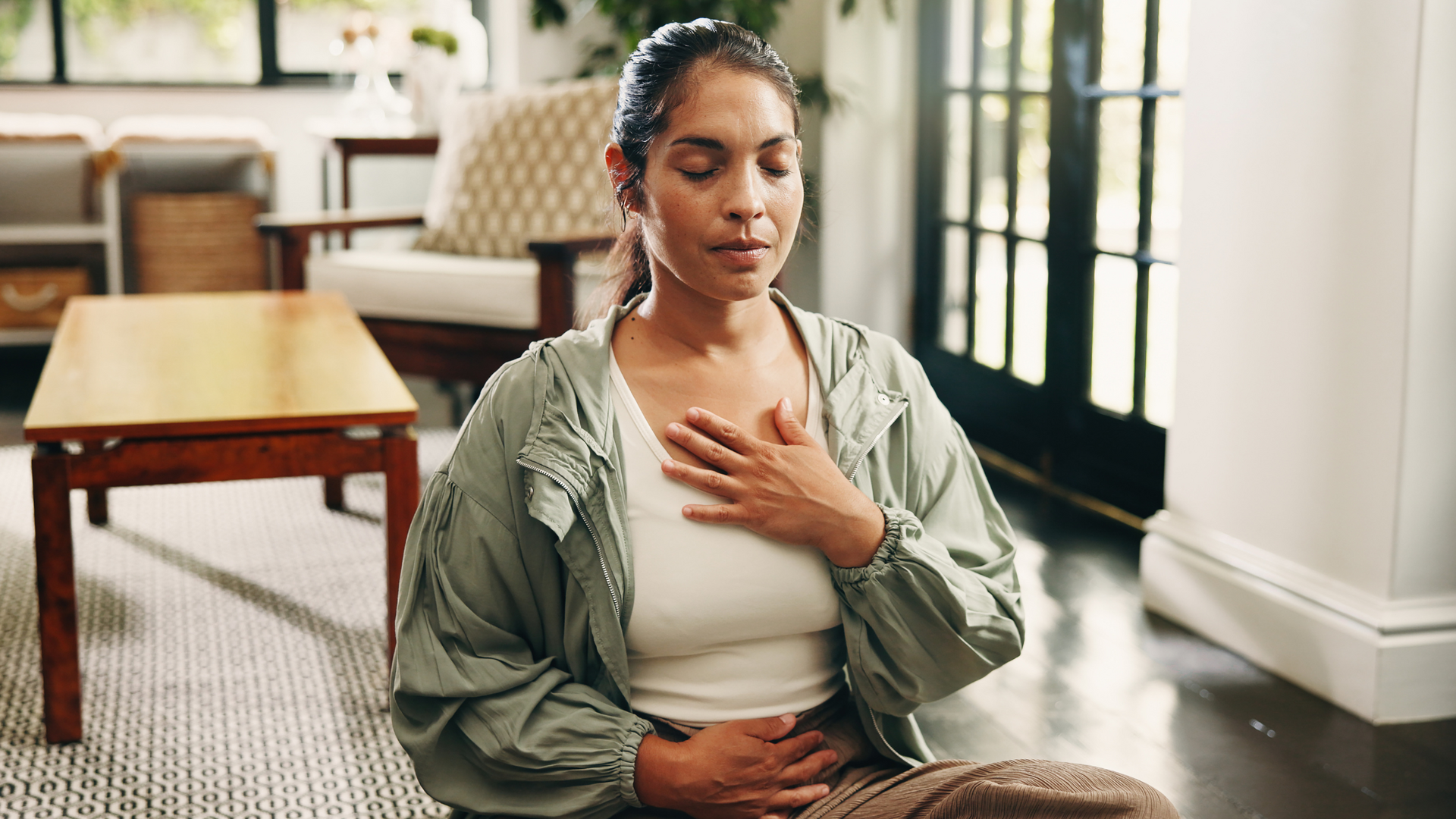 Person practicing breathwork, sitting cross-legged on a yoga mat indoors, eyes closed, hands on knees, focusing on deep, mindful breathing techniques.