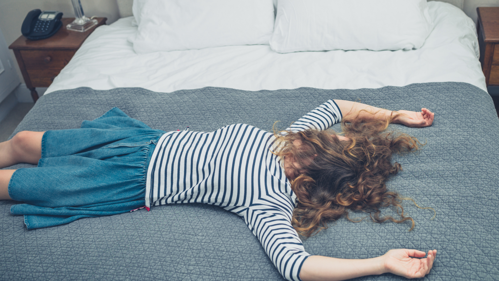A tired woman sits slumped at her desk, eyes closed and head resting on her arms, surrounded by paperwork and an empty coffee cup, looking exhausted.
